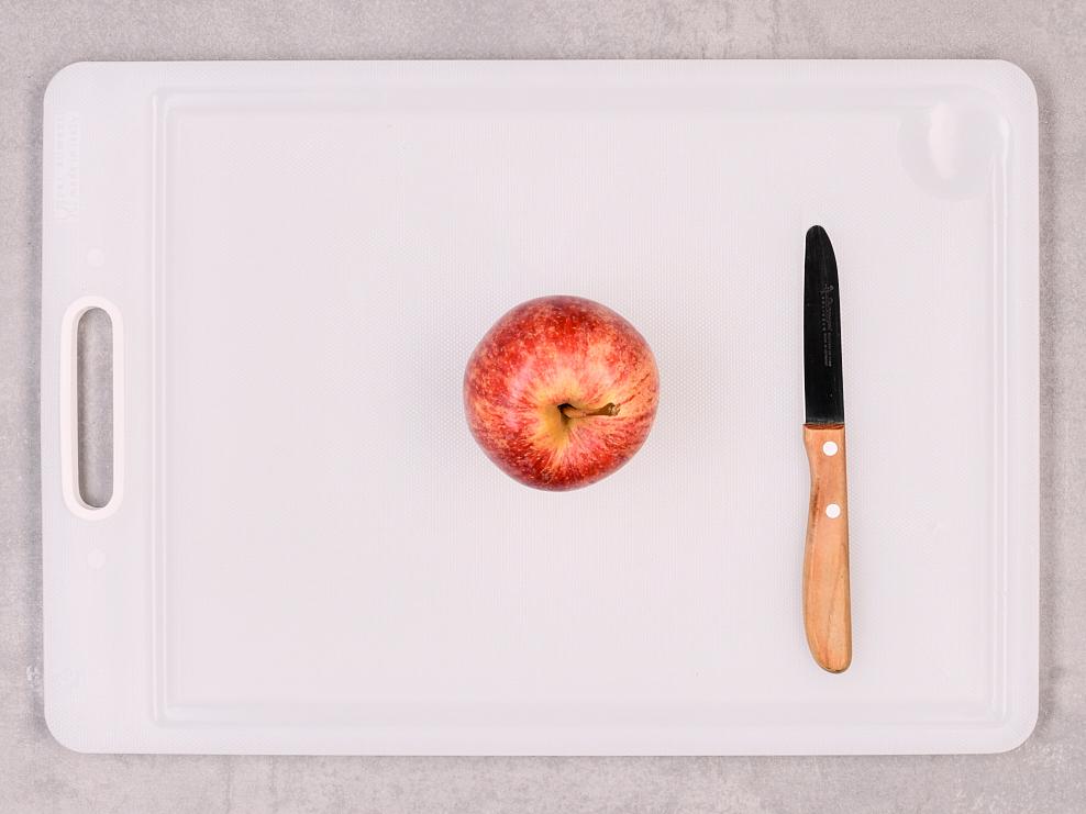 Roter Apfel liegt neben Küchenmesser auf weissem Schneidebrett.