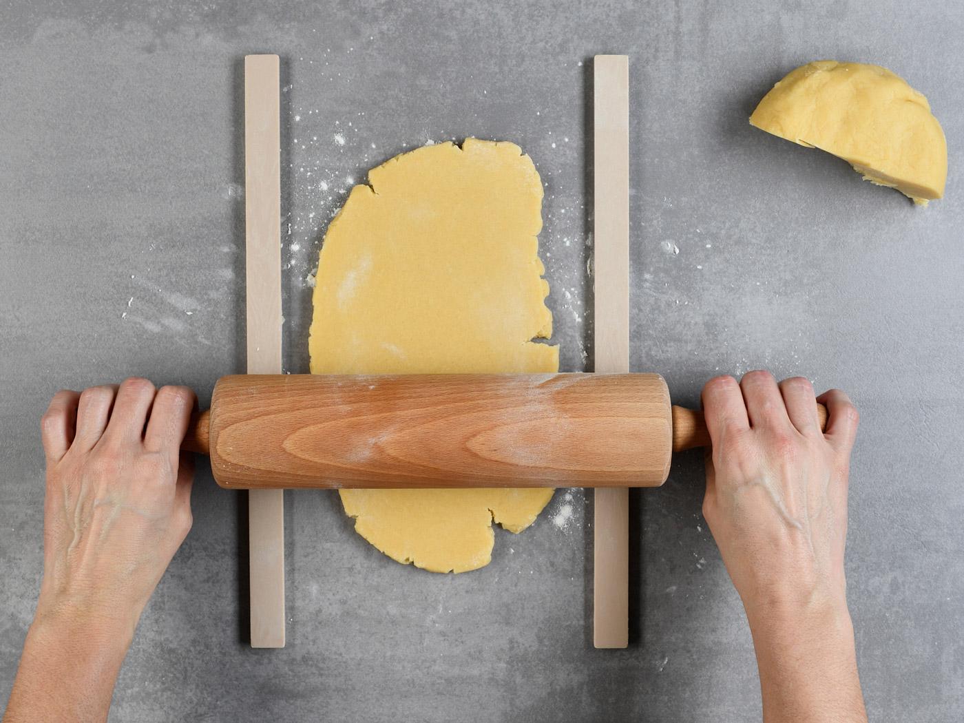 Zwei Hände rollen Teig mit einem Wallholz zwischen Holzleisten aus.