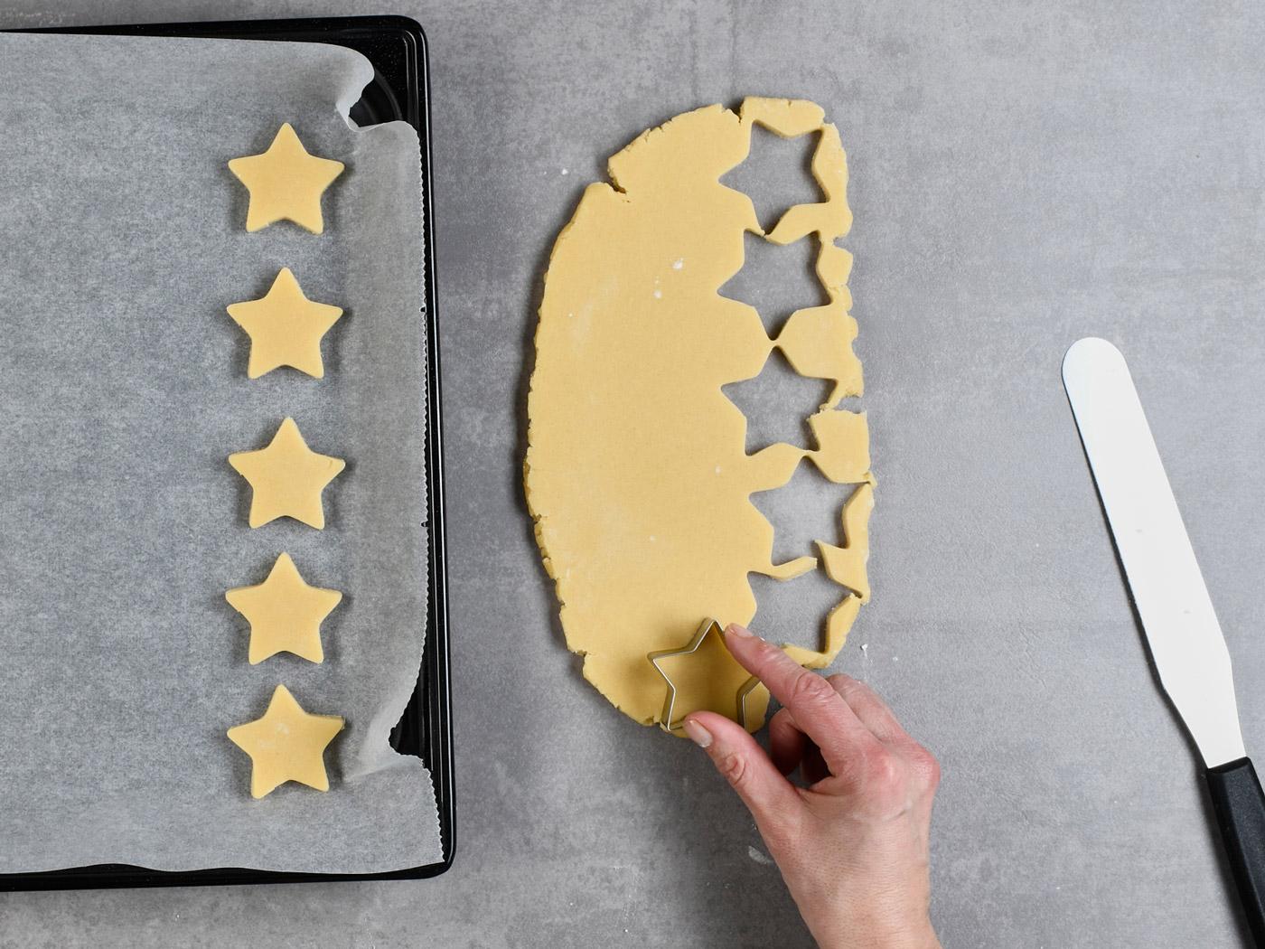 Hand sticht Sterne aus Teig aus, daneben Backblech mit ausgestochenen Sternen.