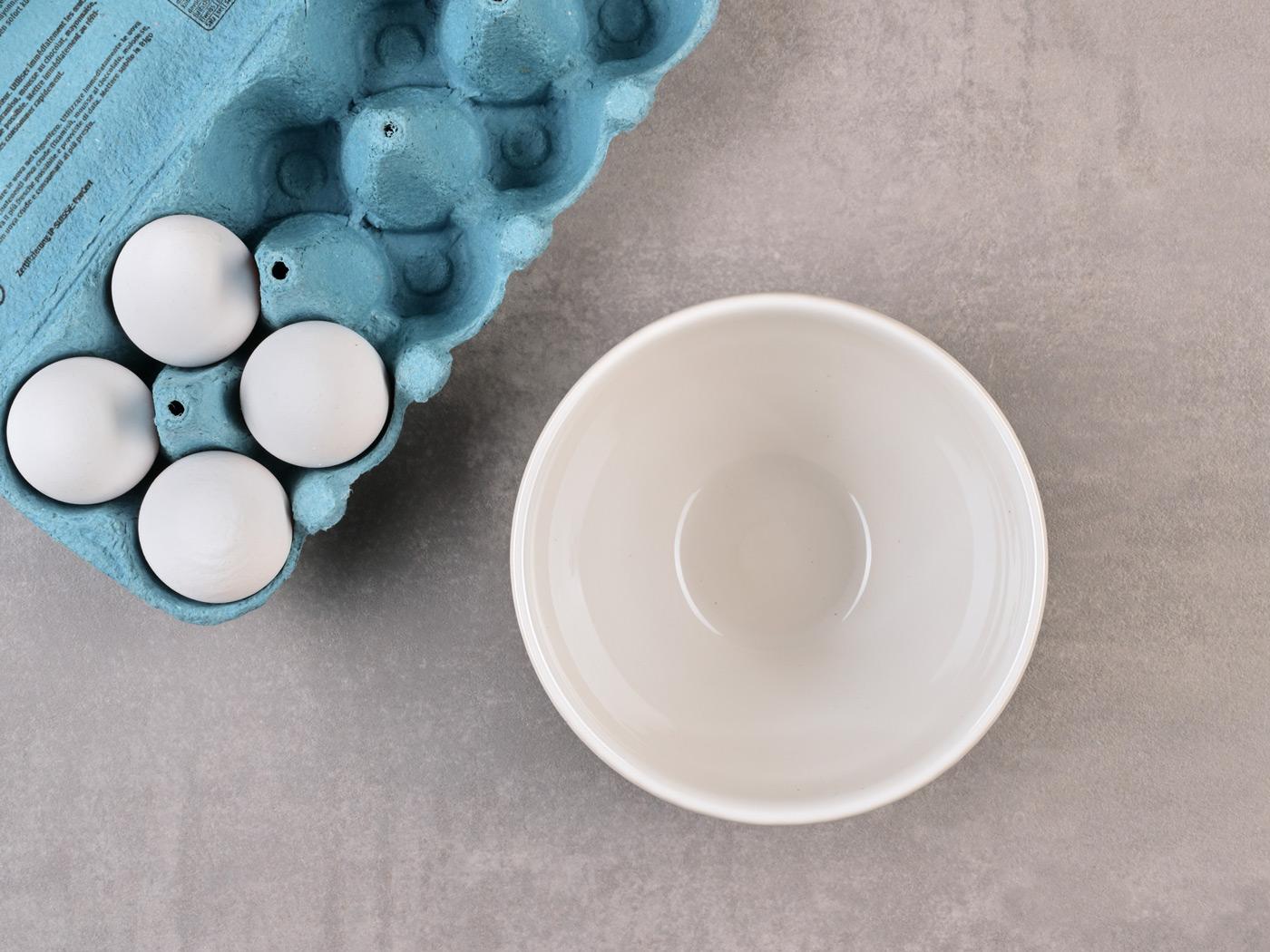 Vier Eier in blauer Schachtel neben leerer weisser Schüssel.
