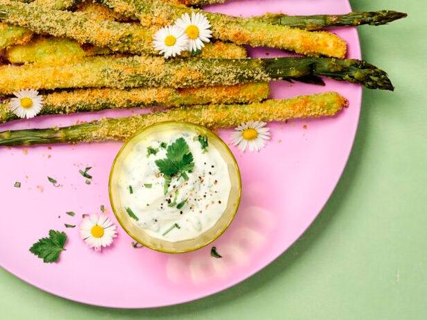 Panierte grüne Spargelstangen mit Dip auf pinkem Teller, mit Kräutern und Gänseblümchen.
