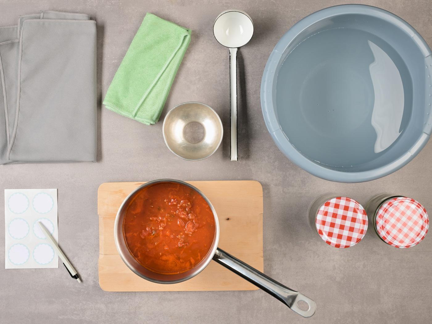 Topf mit Tomatensauce und Einmachutensilien auf Tisch.