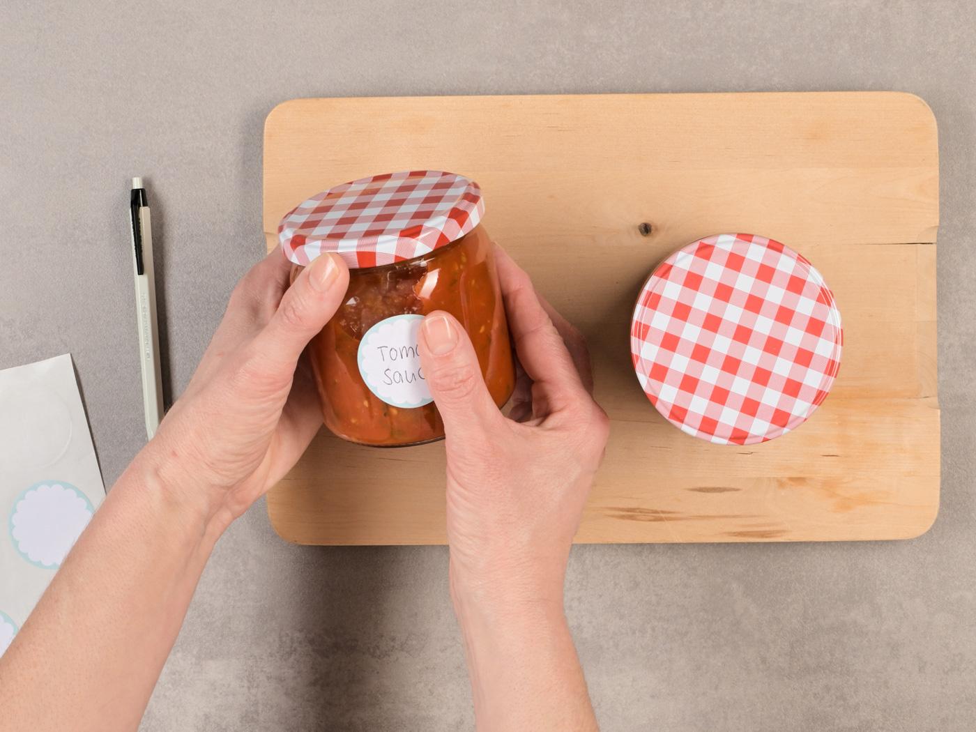 Hände halten ein Glas Tomatensauce mit kariertem Deckel und Etikette.