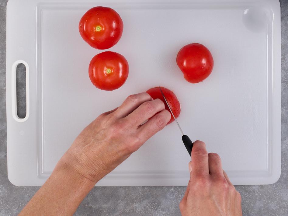 Vier rote Tomaten liegen auf einem Brett, eine wird in Scheiben geschnitten.