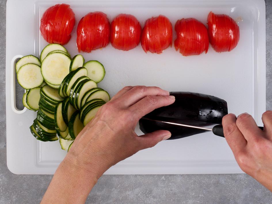 Auf einem Schneidebrett liegen aufgeschnittene Tomaten und Zucchini, eine Aubergine wird geschnitten.