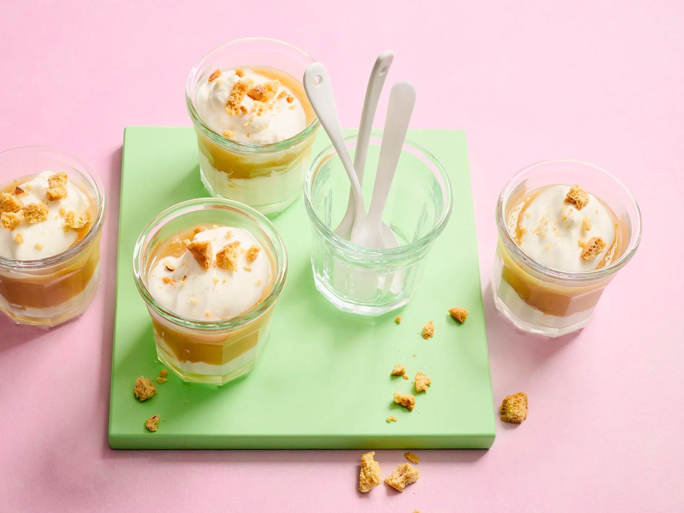 Vier Dessertgläser mit Creme und Rahm, bestreut mit Guetzli-Stücken auf rosa Hintergrund.