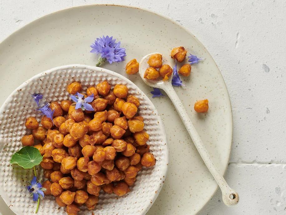 Teller mit gerösteten Kichererbsen und Blüten, daneben ein Löffel mit Kichererbsen.
