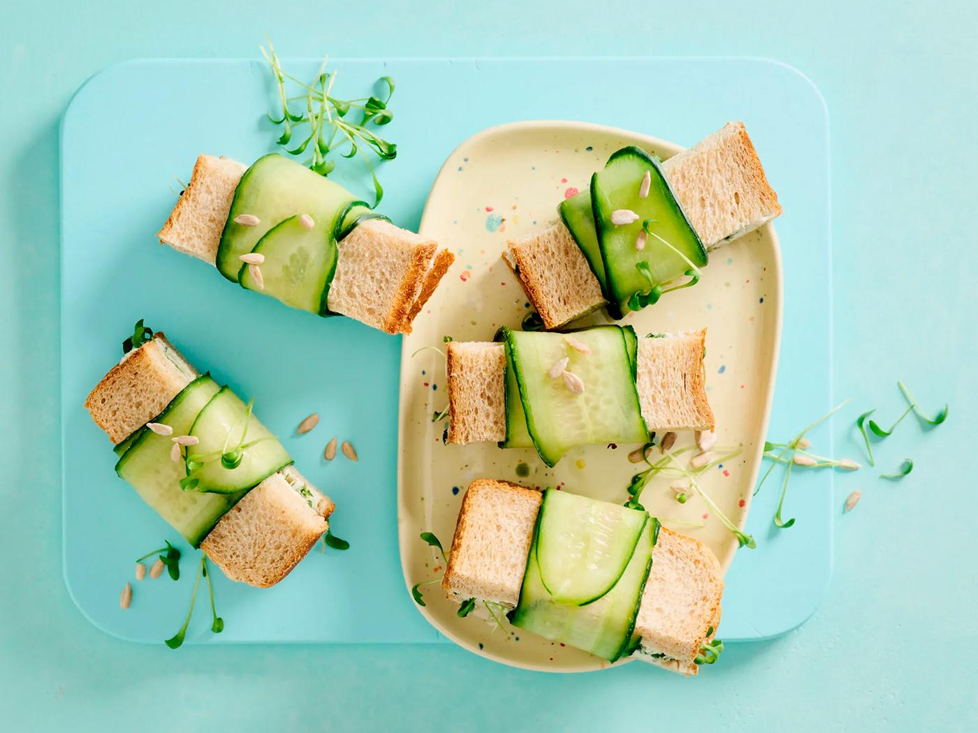 Kleine Sandwiches mit Gurkenscheiben umwickelt, auf hellblauen Tellern angerichtet.