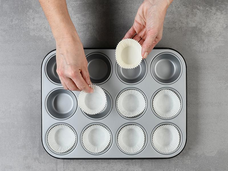 Hände setzen weisse Papierförmchen in ein Muffinblech ein.