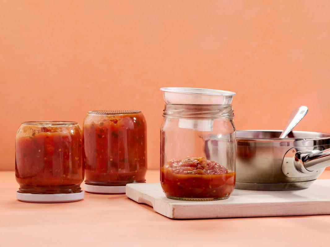 Eingekochte Tomatensauce in drei Einmachgläsern neben einem Kochtopf.